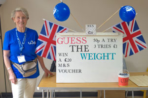 A woman in a blue top with the u3a logo next to a white board that says, 'guess the weight' and union jack flags