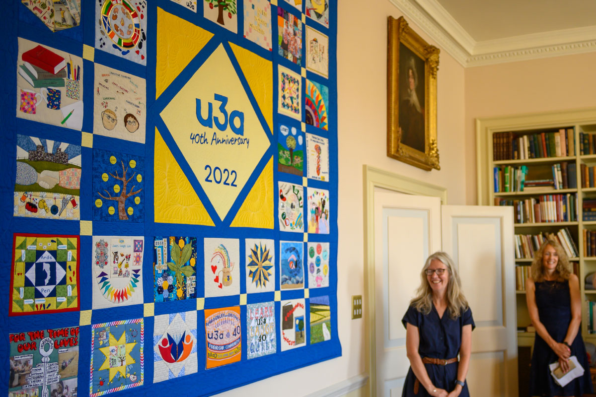 A close up of a quilt made up of colourful blocks on a blue background. In the middle of the quilt is a yellow square, and in that a diamond, with the words, "u3a 40th Anniversary 2022." Two women are standing next to the quilt, looking at it and smiling.
