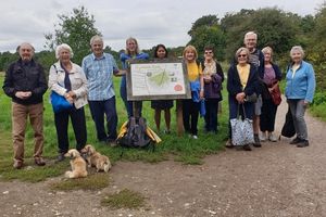 Around 12 older adults are standing on a green field next to a trail map. They have two small dogs with them.