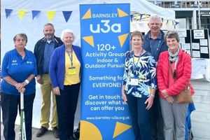 Six older adults stand next to a big blue stand up banner that reads 'Barnsley u3a 120+ Activity groups Indoor & Outdoor Pursuits, Something for EVERYONE!" 