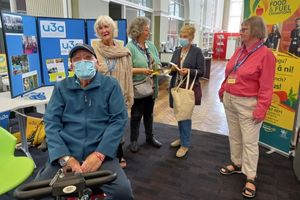A man wearing a blue face mask is sitting in a library, behind is a stand that says u3a and has pictures on it. Three women are standing behind him, chatting.