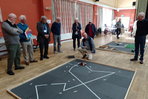 A group of men are standing around a black board on the floor with white lines drawn on it. On the far end of the board is a small robot, and a man is kneeling by it smiling.