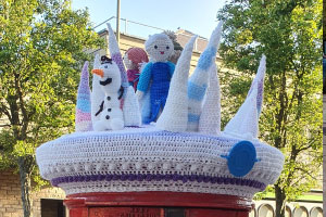 a postbox with a knitted covering consisting of icicles, a woman wearing a blue dress and snowman