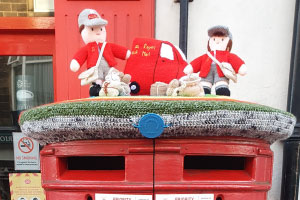 a postbox with a knitted covering consisting of a knitted postman, postwoman and lorry