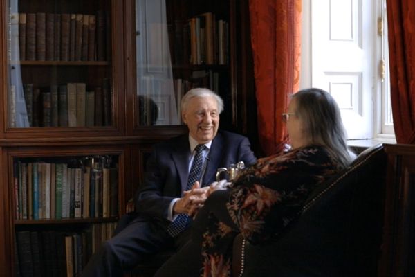 A man and woman are talking in a library. The man is facing the woman and is smiling. The woman's back is to us.