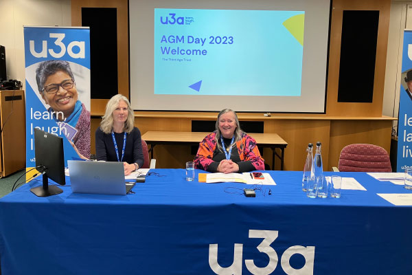 CEO of the Third Age Trust, Sam Mauger and Chair of the Third Age Trust, Liz Thackray sitting behind a table with a blue u3a table cloth on it. Behind them are two u3a flag banners and a PowerPoint presentation that reads, "AGM Day 2023 Welcome"