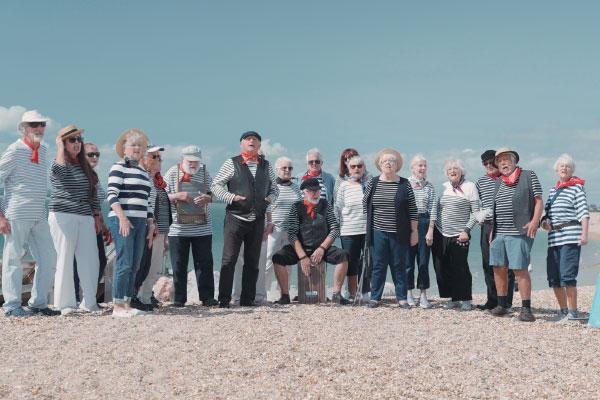 A group of people, all wearing stripy shirts, standing on a beach, singing
