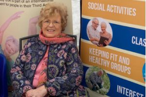 A woman in a pink top and floral jacket sitting down next to a blue and yellow poster