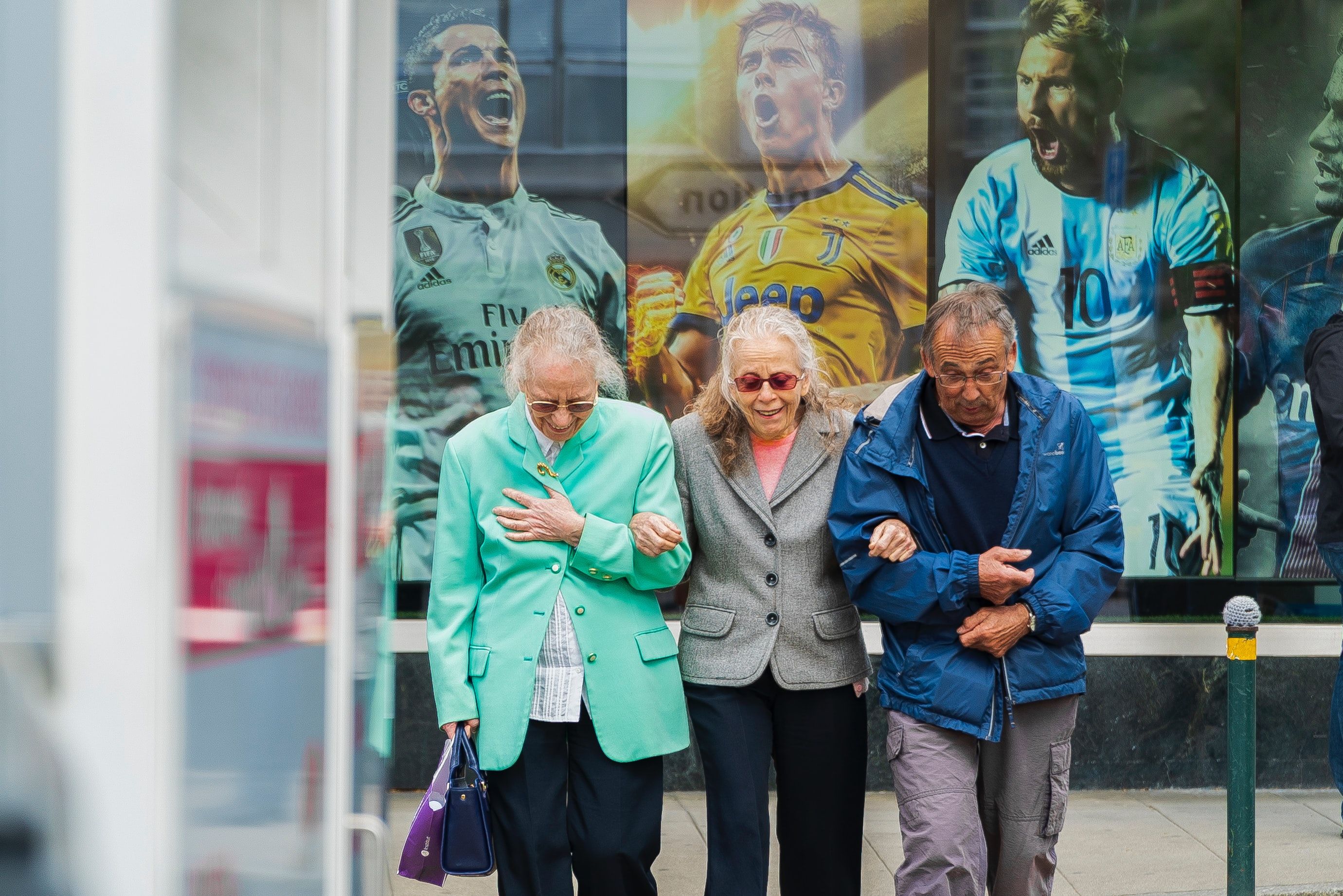 Three older people linking arms smiling and walking away from a poster of 3 male football players