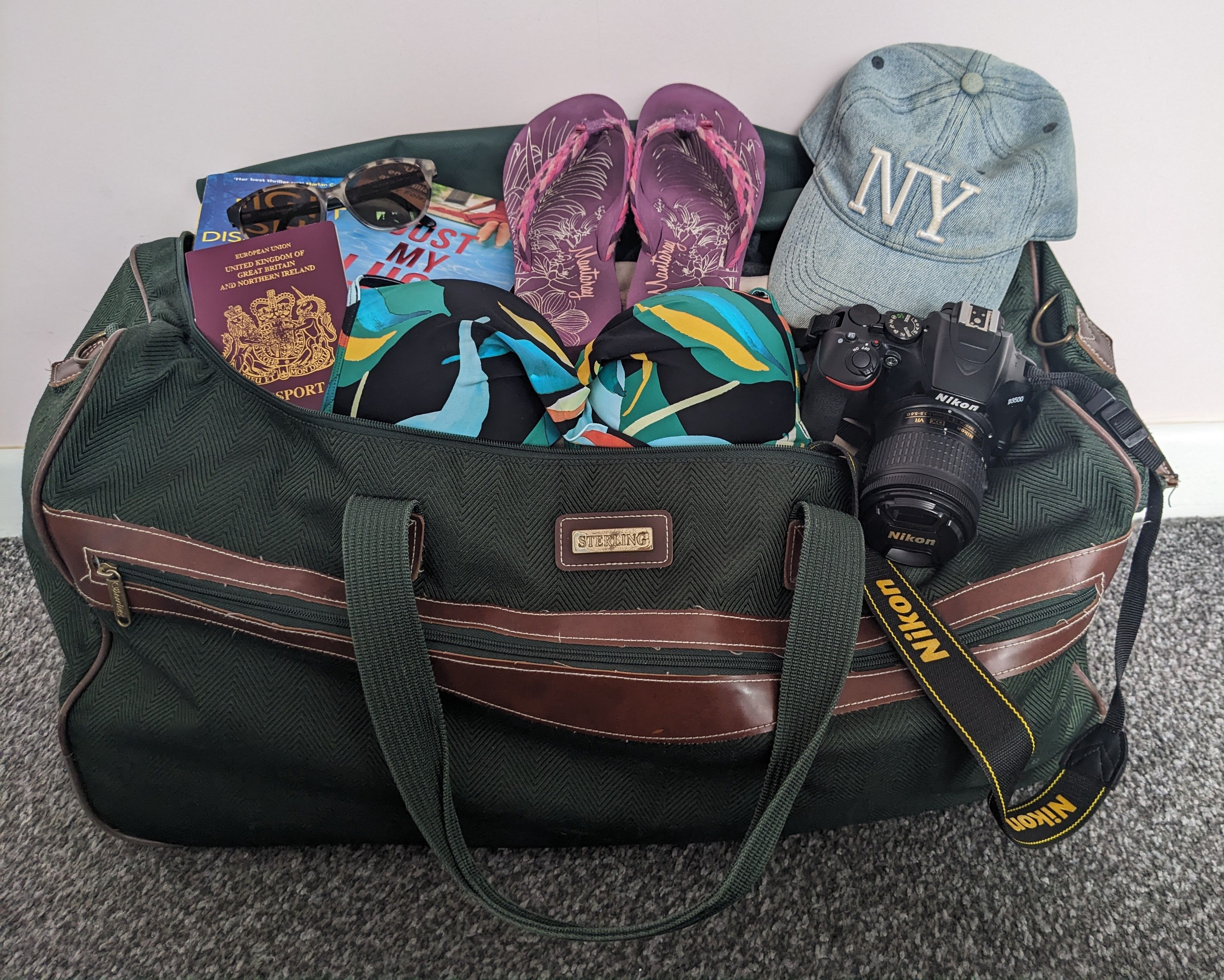 a green and brown holdall bag packed full of summer holiday essentials - passport, flip flops, sunhat, sunglasses, camera etc