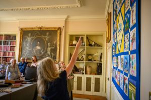 A room with book cases and a painting in the background. On the wall in the foreground right is a colourful quilt made up of square blocks which are all different. In the background of the room, several women look up at the quilt; in the foreground, two women are looking and pointing at the quilt.