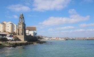 buildings on the left and the blue sea on the right, the sky is blue with small clouds