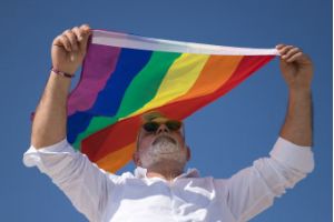 A man with a grey beard wearing a white shirt holding up a pride flag against a blue sky