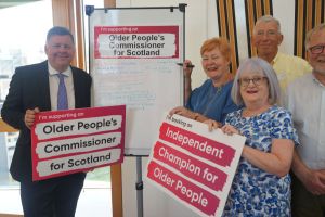 A group of people standing next to a whiteboard. The Whiteboard says "Commissioner for Older People' and underneath there are many signatures. A woman with ginger hair is holding a pen, about to sign. The man on the left hand side of the whiteboard is holding a banner saying, "I support a Commissioner for Older People and Ageing." On the other side of the whiteboard, a woman is holding a banner saying, "Independent Champion for Older People."