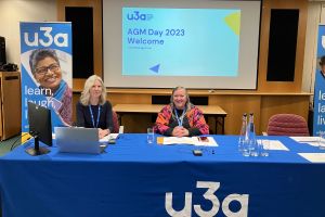 CEO of the Third Age Trust, Sam Mauger and Chair of the Third Age Trust, Liz Thackray sitting behind a table with a blue u3a table cloth on it. Behind them are two u3a flag banners and a PowerPoint presentation that reads, "AGM Day 2023 Welcome"
