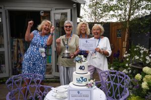 Four women are holding a trophy and a medal, they look ecstatic and are smiling and cheering. They are stood in a garden, behind a table with a cake that says '40 u3a Southport'