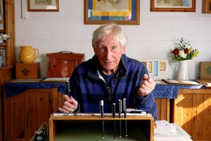 A man in a blue and black striped jumper is sitting at a desk in his home. The desk is slightly slanted and there are pens on it. The man is holding one pen above a piece of paper with ink on it, and is looking directly at the camera.